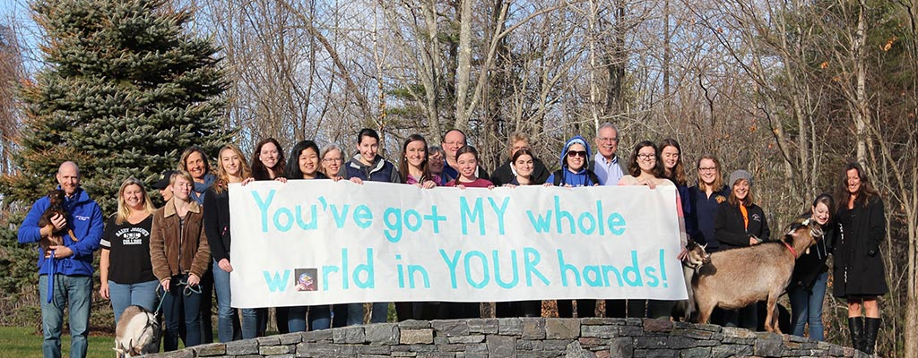 A group of students and goats stand outdoors, holding a large banner that reads, "You've got MY whole world in YOUR hands!" as part of a hashtag campaign for climate change awareness. Saint Joseph's College of Maine