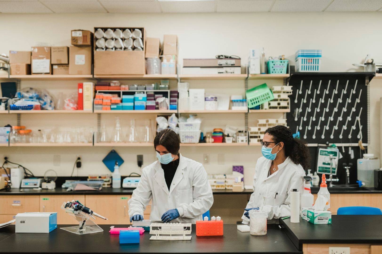 Two scientists wearing lab coats and protective masks work in a laboratory resembling those found at Saint Joseph's College of Maine. The lab, equipped with various scientific instruments and supplies on shelves and countertops, reflects the high standards of the Doctor of Chiropractic Program. Saint Joseph's College of Maine
