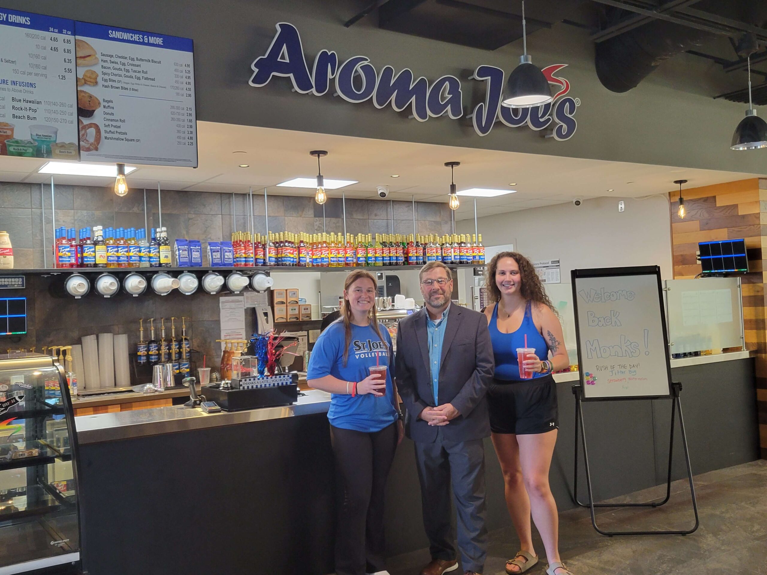 Three people stand in front of an Aroma Joe's coffee shop counter at the bustling SJC campus. Two women hold drinks while a man in a suit stands between them. A menu and various counter items are visible in the background, capturing the lively atmosphere. Saint Joseph's College of Maine