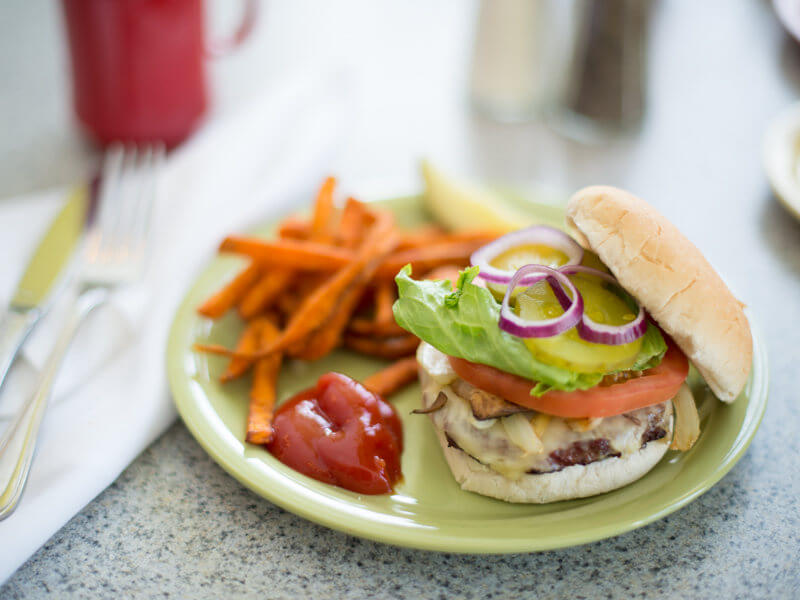 A hamburger with lettuce, tomato, pickles, and red onion sits open-faced on a plate with a side of sweet potato fries and a dollop of ketchup. Fork and knife are placed on a napkin beside the plate, showcasing one of our premium dining services food options. Saint Joseph's College of Maine