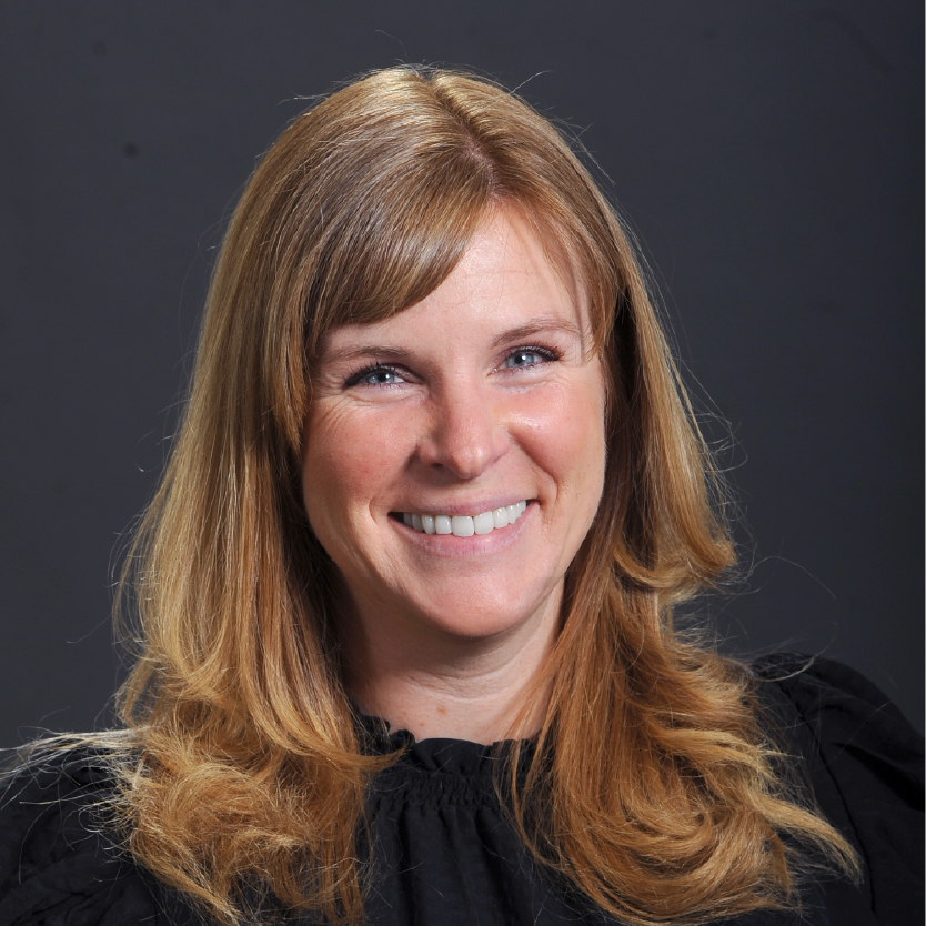 Smiling woman with long blonde hair, wearing a black top, posed in front of a dark gray background. Ashley Shamos, NNP, APRN, exudes professionalism and warmth in this portrait. Saint Joseph's College of Maine