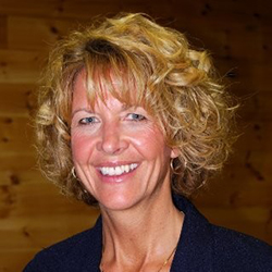A woman with curly blonde hair, smiling, is wearing a dark jacket. The background features wooden paneling, evoking the cozy charm of a Maine college. Saint Joseph's College of Maine
