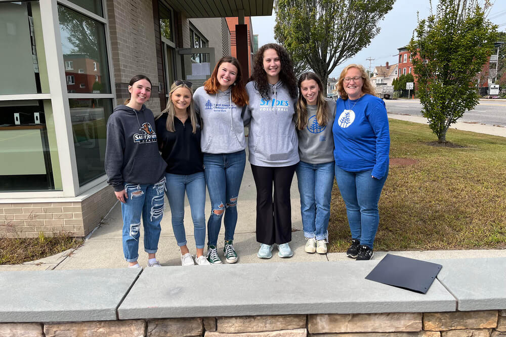Community Service 3 Six women are standing in a line outdoors, smiling at the camera. They are casually dressed, wearing jeans and a mix of sweatshirts and hoodies. They're next to a building with a sidewalk and grassy area, possibly having just finished some Community Service at Mercy Center. Saint Joseph's College of Maine