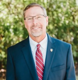 Joseph L. Cassidy, in a navy suit, red striped tie, and glasses, stands outdoors in front of green foliage, smiling at the camera. Saint Joseph's College of Maine