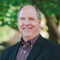 Smiling older man with short gray hair wearing a dark blazer and a checkered shirt, standing outdoors in front of green, leafy trees, celebrating his college degree earned in Maine. Saint Joseph's College of Maine