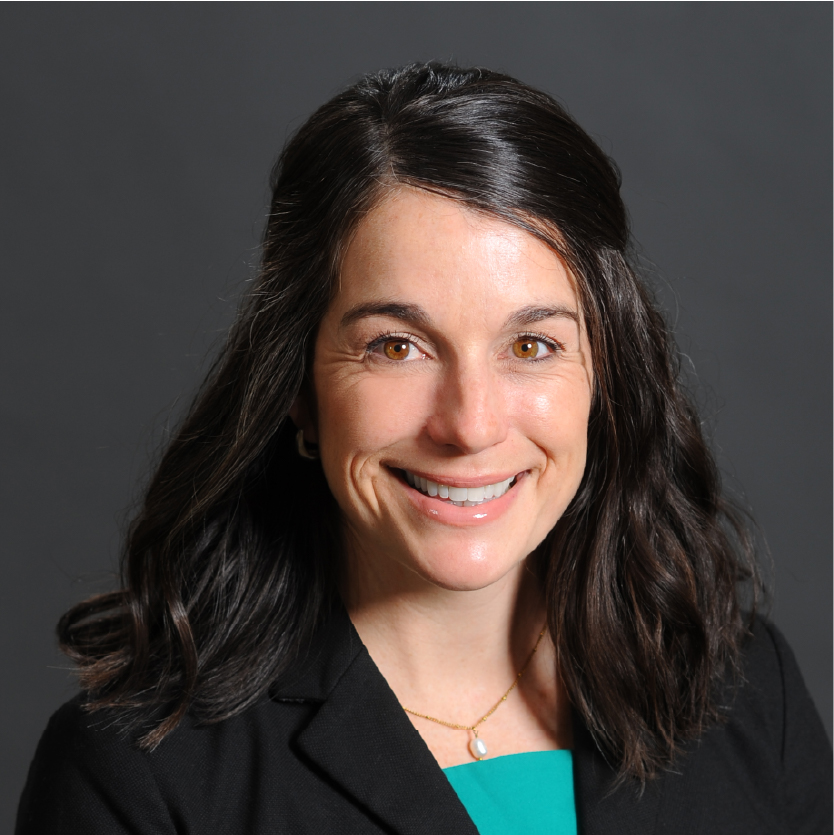Smiling woman with wavy dark hair, wearing a black blazer over a green top and a gold necklace, in front of a plain dark gray background. Lindsay Snyder, AGPCNP-BC, exudes professionalism and warmth in this portrait. Saint Joseph's College of Maine