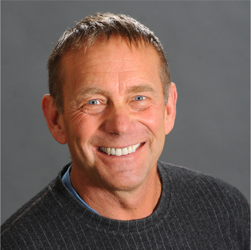 Smiling man with short brown hair, identified as Mark A. Green, wears a dark ribbed sweater over a blue shirt, posing in front of a plain gray background. Saint Joseph's College of Maine