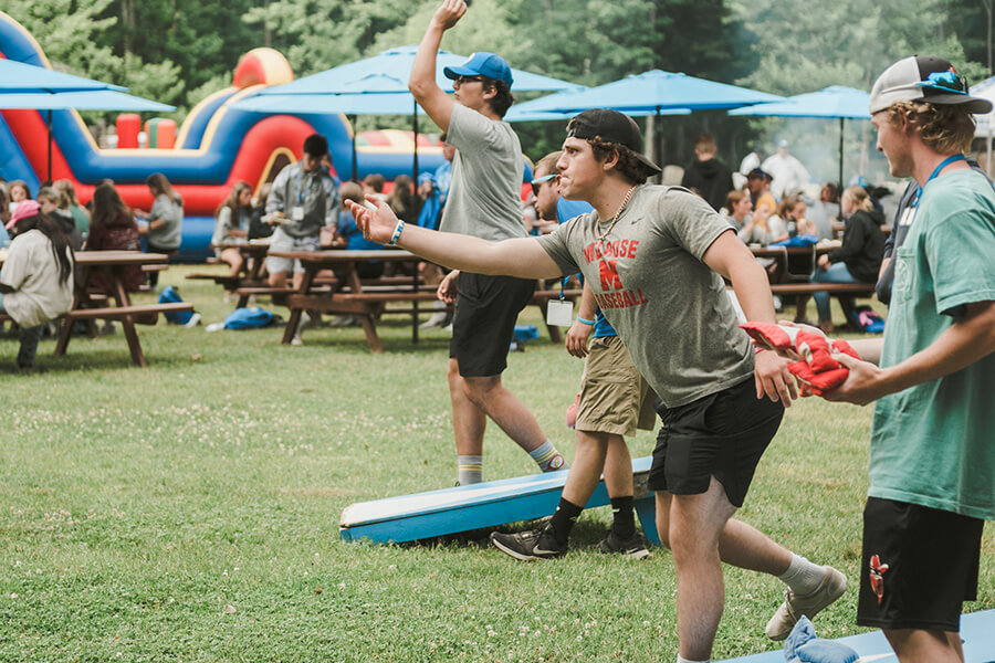 People enjoying a bean bag toss game at an outdoor event, where picnic tables and umbrellas dot the background. The lively scene reflects vibrant student life, with an inflatable obstacle course seen in the distance. Saint Joseph's College of Maine