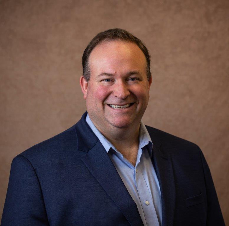 A man wearing a navy blue blazer and light blue shirt smiles at the camera in front of a plain brown background, proudly showing off his college degree from Maine. Saint Joseph's College of Maine