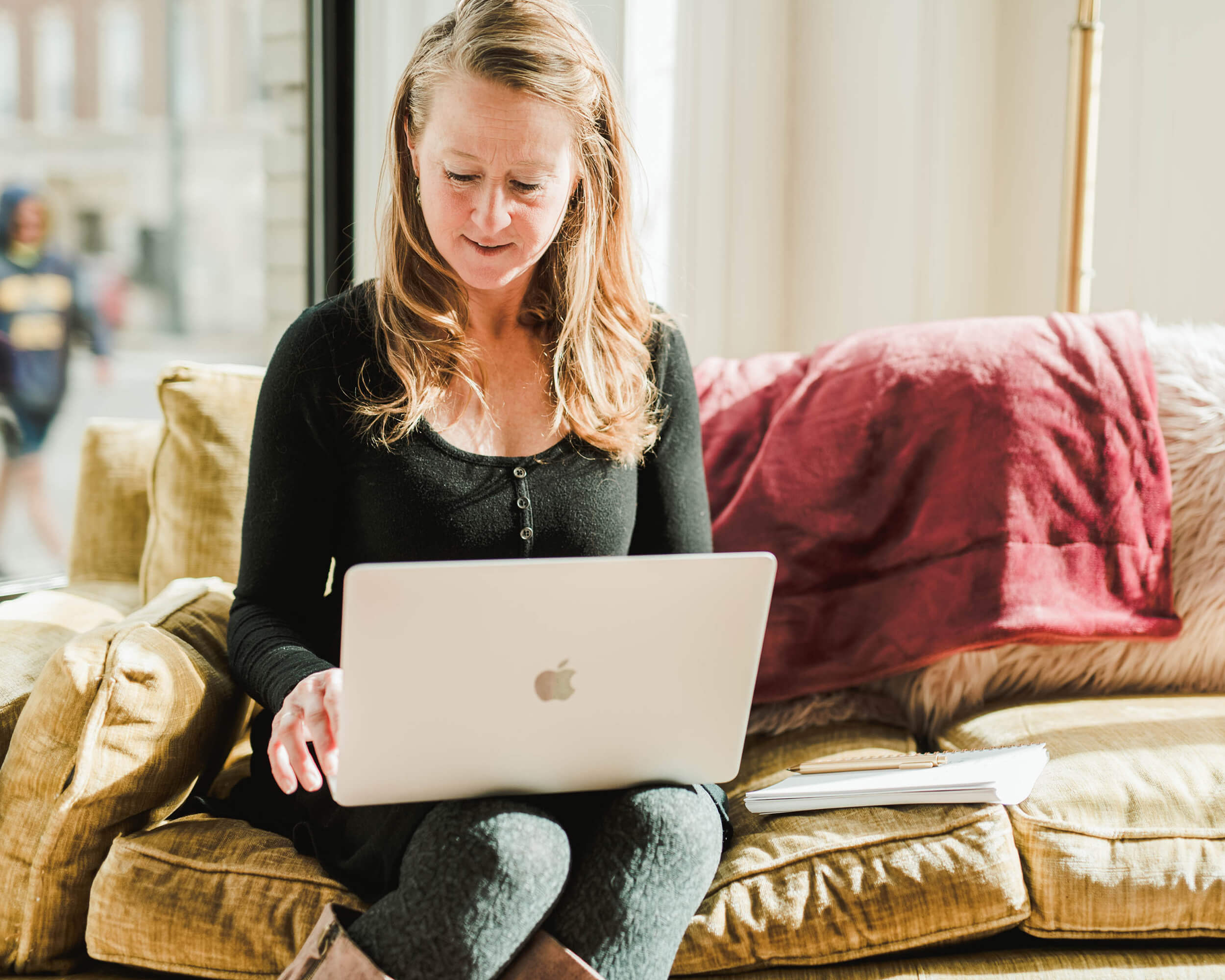 A woman with long hair works on a laptop while sitting on a yellow couch beside a window, her notes for an online graduate health administration degree scattered nearby, along with a red blanket. Saint Joseph's College of Maine