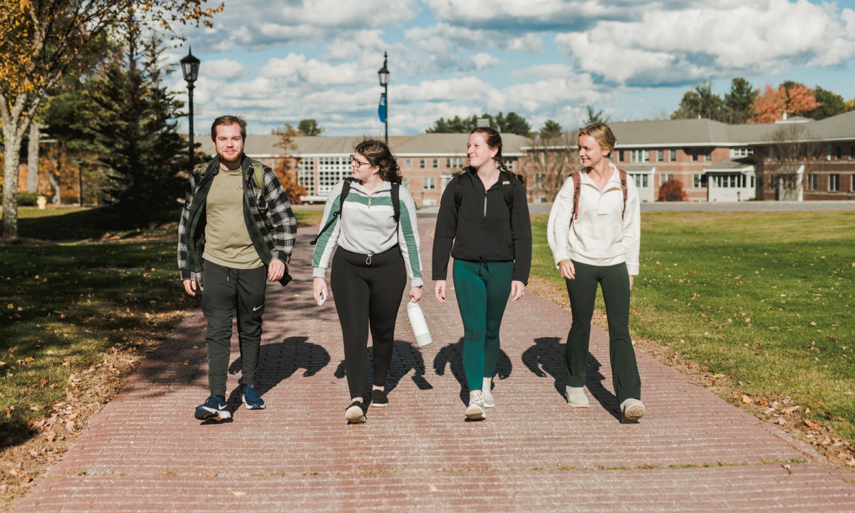 Business Programs 1 Four people walk side by side on a brick pathway through a green campus area with trees and a building in the background. Saint Joseph's College of Maine