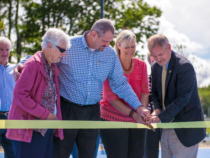 Four people participate in a ribbon-cutting ceremony outdoors at Saint Joseph's College of Maine, using large scissors to cut a yellow ribbon. Trees and a partly cloudy sky are visible in the background. Saint Joseph's College of Maine