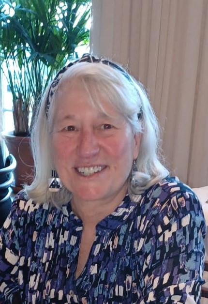 A smiling older woman with white hair, wearing a patterned blouse and headband, sits indoors with a leafy green plant in the background, proudly reflecting on her college degree from Maine. Saint Joseph's College of Maine
