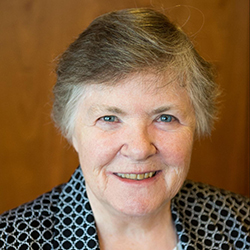 An older woman with short gray hair, light eyes, and a friendly smile, wearing a black and white patterned jacket. The plain brown background hints at her Maine college roots and the pride she takes in her degree. Saint Joseph's College of Maine