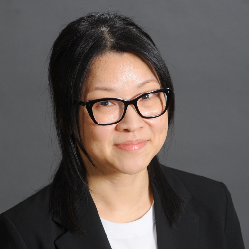 Woman with long black hair and glasses, wearing a black blazer over a white top, smiles slightly at the camera. Yi Jin Kim against a gray background. Saint Joseph's College of Maine