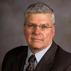 A middle-aged man with short gray hair and glasses, wearing a dark suit, striped shirt, and patterned tie, poses against a neutral background—his confidence reflecting his college degree earned in Maine. Saint Joseph's College of Maine