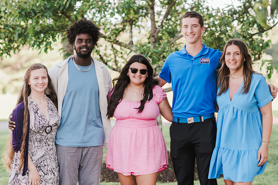 Five people standing together outdoors, smiling at the camera. The three women, possibly parents, are in dresses of different styles while the two men are wearing casual clothing. They are standing in front of green foliage. Saint Joseph's College of Maine