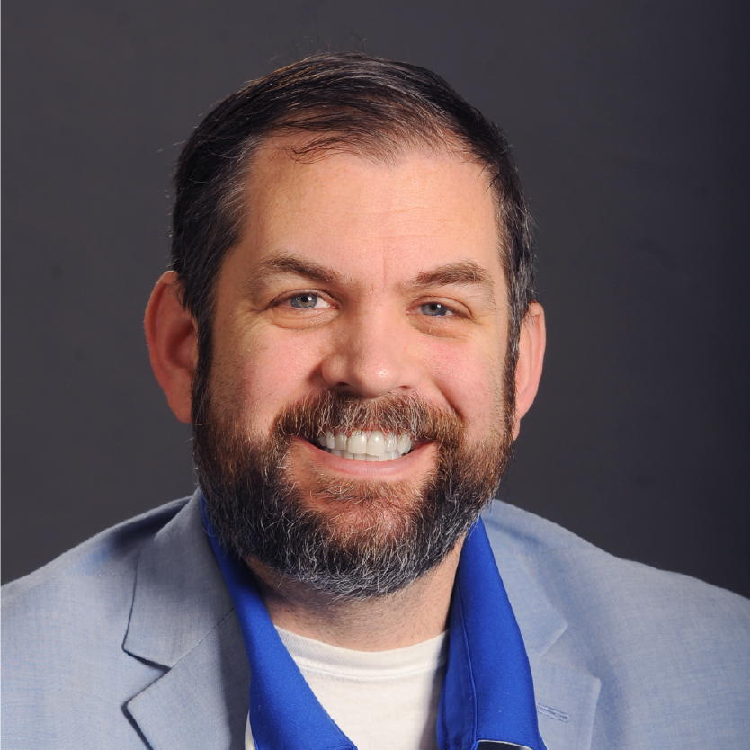 Smiling man with short dark hair and a full beard, wearing a light blue blazer, blue collared shirt, and white undershirt, against a plain dark background—Christopher Woodside. Saint Joseph's College of Maine