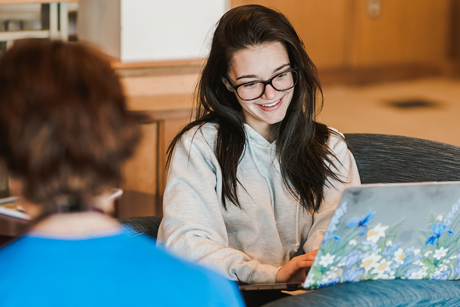 A person with long hair and glasses is sitting and smiling while using a laptop with a floral design on the cover, possibly working on a redesign. Another person ina blue shirt is visible in the foreground, looking like one of their parents. Saint Joseph's College of Maine