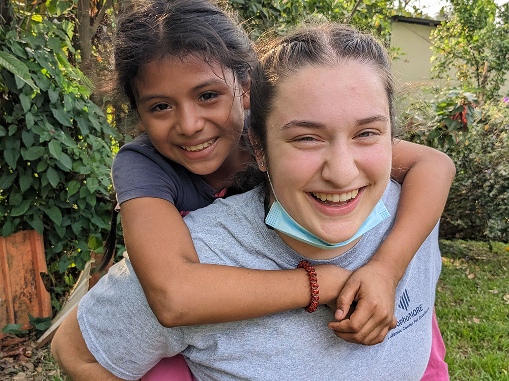 Service Trips 6 Two smiling girls outdoors, with the younger one piggybacking on the older one. The older girl has a face mask hanging below her chin. They are surrounded by greenery, capturing a joyful moment during their Guatemala service trip. Saint Joseph's College of Maine