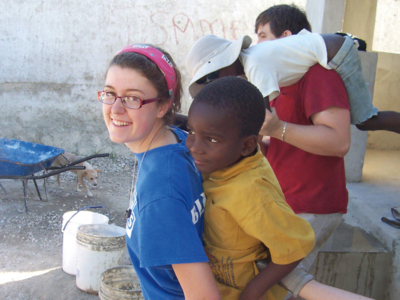 Service Trips 8 A smiling young woman carries a child on her back outdoors, perhaps enjoying a break from college. Another person nearby carries a second child over their shoulders, with buckets and a wheelbarrow visible in the background. Saint Joseph's College of Maine