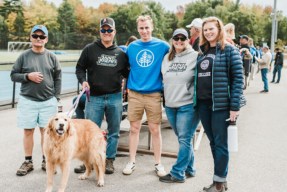 A group of five people, including two parents, stands outdoors with a dog. Some are wearing Saint Joseph's College apparel. Trees and a sports field are visible in the background. Saint Joseph's College of Maine