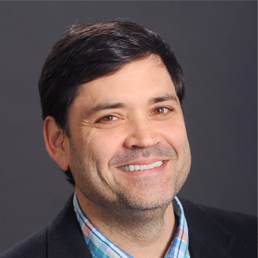 Smiling man with short dark hair, wearing a blue plaid shirt and black blazer, posed against a plain dark background. Saint Joseph's College of Maine