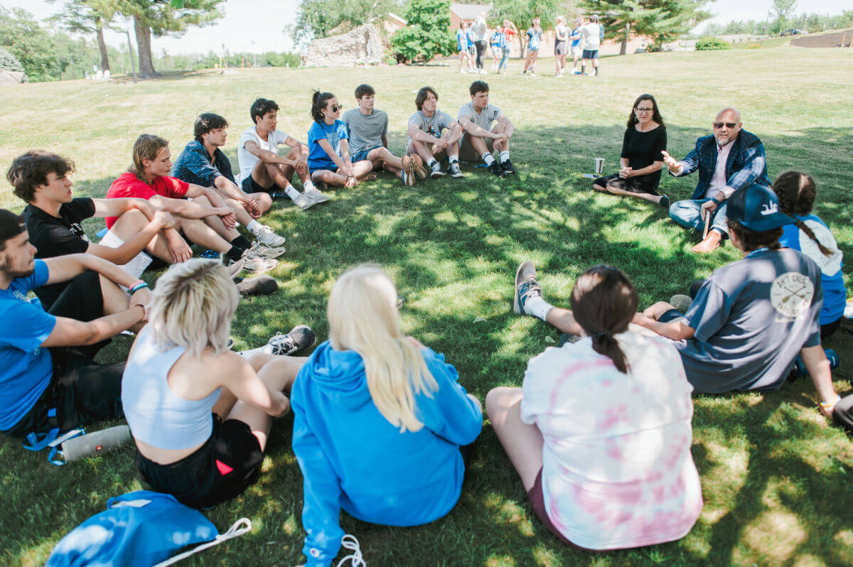 Community Leadership 1 A group of teens and two adults sit in a circle on grass outdoors, listening and talking—perhaps discussing college plans or a future degree. Some people walk on a path in the background. It is sunny with Maine's trees providing shade. Saint Joseph's College of Maine
