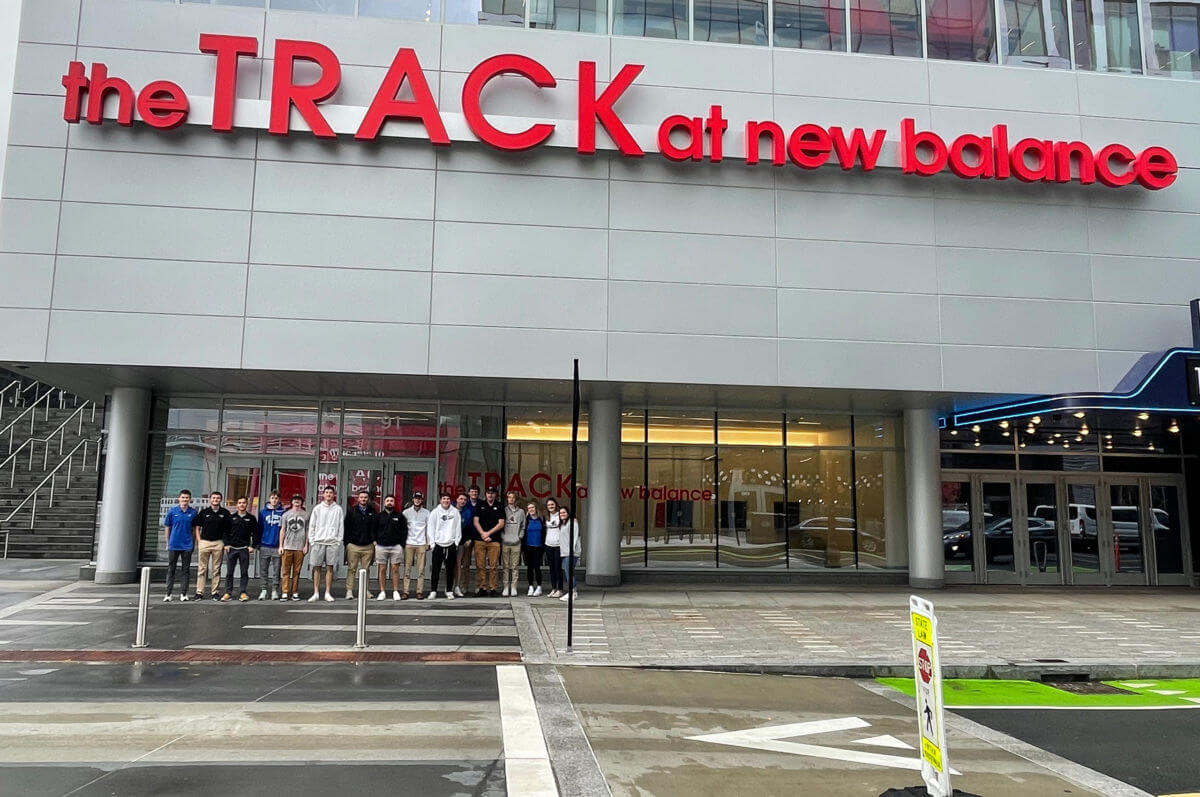 A group of people stands in front of a building with the sign "the TRACK at new balance." The building exterior is modern with glass windows, showcasing a space that complements sport and exercise science programs. Saint Joseph's College of Maine