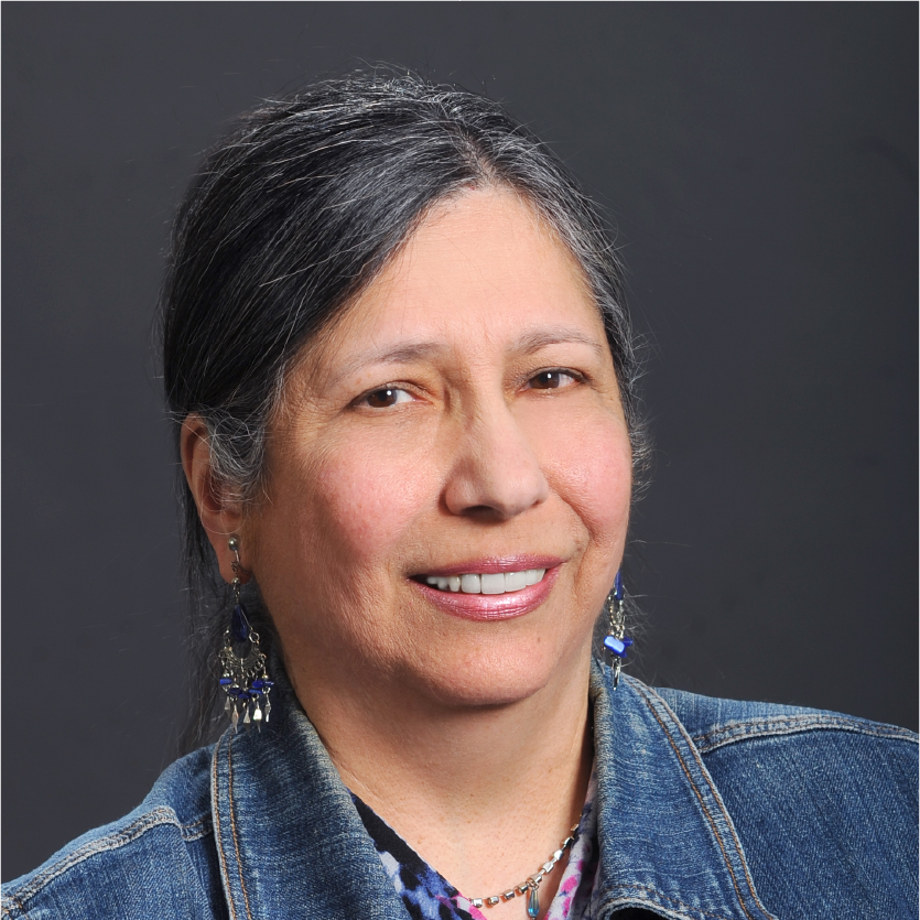 Nina B. Eduljee, a woman with gray and black hair, smiles gently. She wears a denim jacket, patterned shirt, and beaded earrings, and is posed against a plain dark background. Saint Joseph's College of Maine