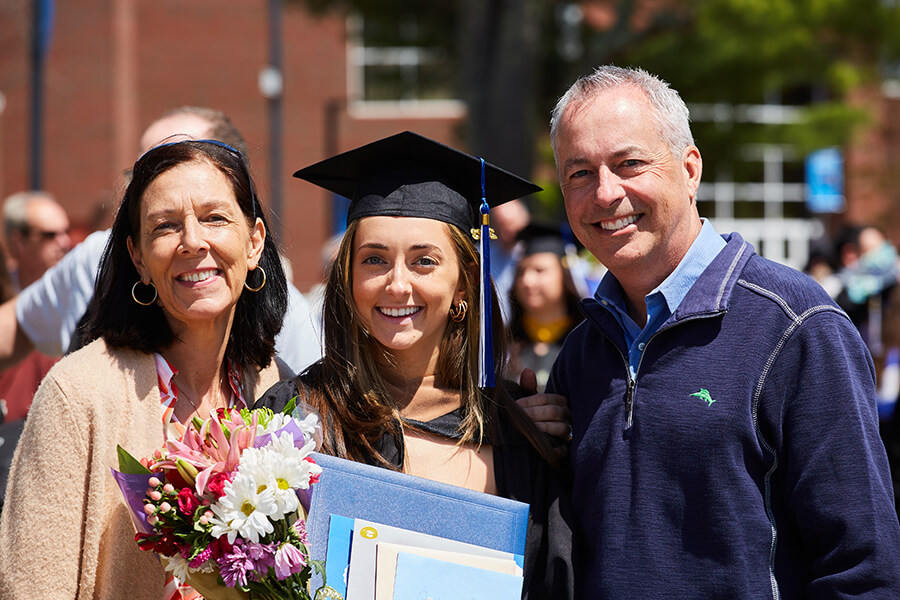 A graduate in a cap and gown stands between proud parents—an older woman holding flowers and an older man—all smiling. Saint Joseph's College of Maine