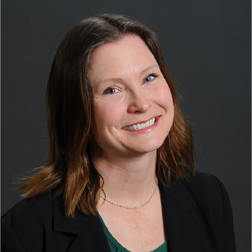 Patricia Waters, a woman with shoulder-length brown hair, smiles at the camera. She is wearing a black blazer over a green top, with a thin necklace. The background is solid dark gray. Saint Joseph's College of Maine