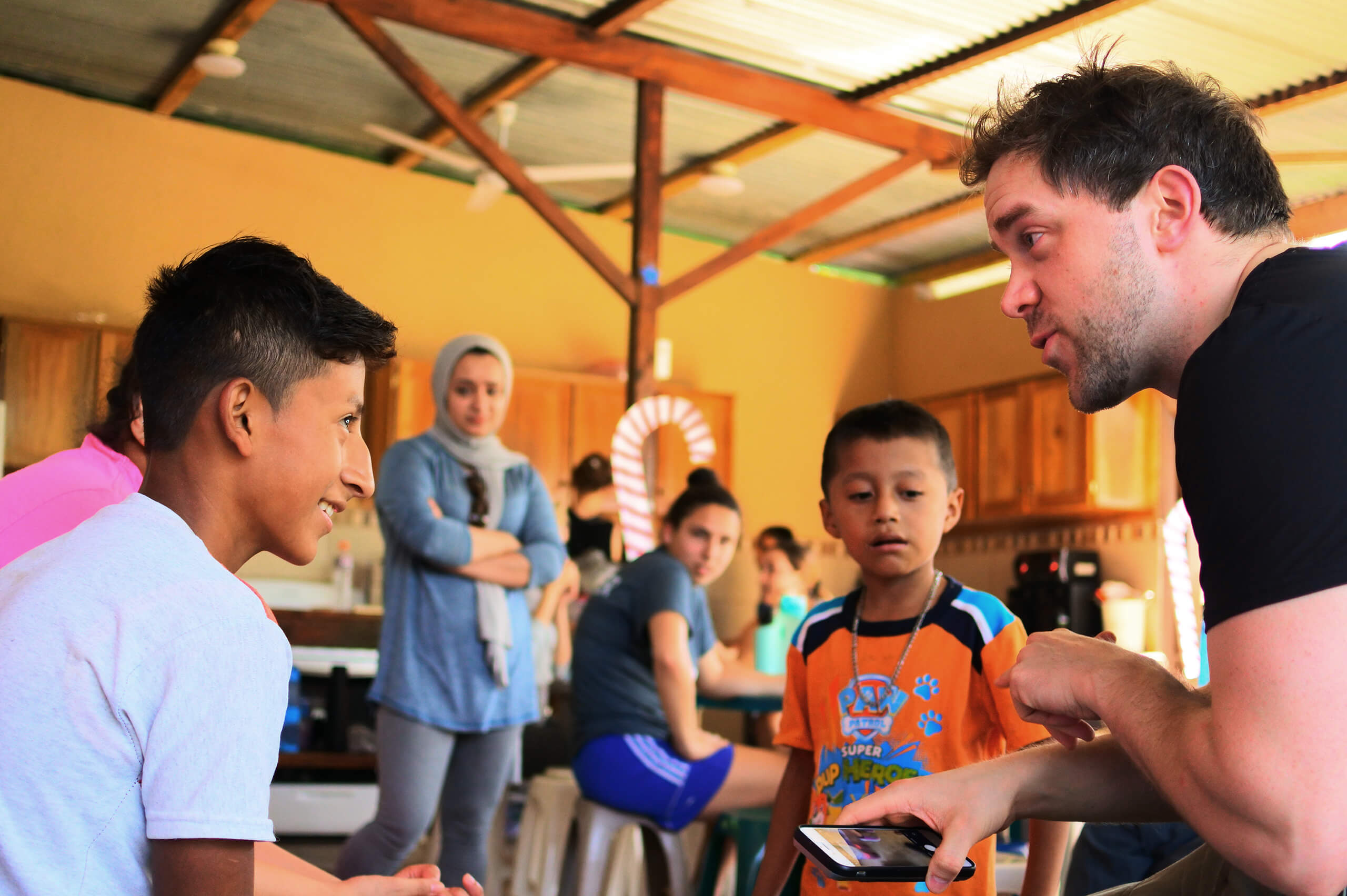Theology Programs 1 A man holding a smartphone talks animatedly with two boys in a lively indoor setting, possibly at a college event in Maine, while others watch and interact in the background, suggesting a casual, friendly gathering. Saint Joseph's College of Maine