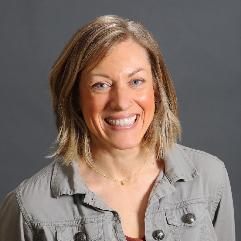A woman with short, light brown hair smiles at the camera. She is wearing a gray button-up shirt and a gold necklace, with a neutral gray background behind her. Saint Joseph's College of Maine