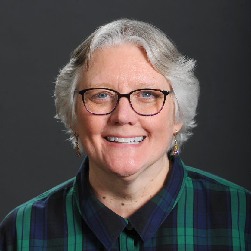 Smiling woman, Wendy Galgan, with short gray hair and glasses, wearing a blue and green plaid shirt, in front of a plain dark background. Saint Joseph's College of Maine