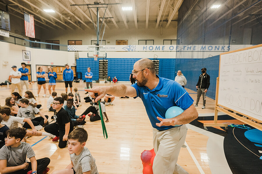 A coach wearing a blue shirt speaks passionately while pointing in a gymnasium filled with seated children and adults. A whiteboard with writing, outlining Sport and Exercise Science undergraduate programs in Maine, is visible in the background. Saint Joseph's College of Maine
