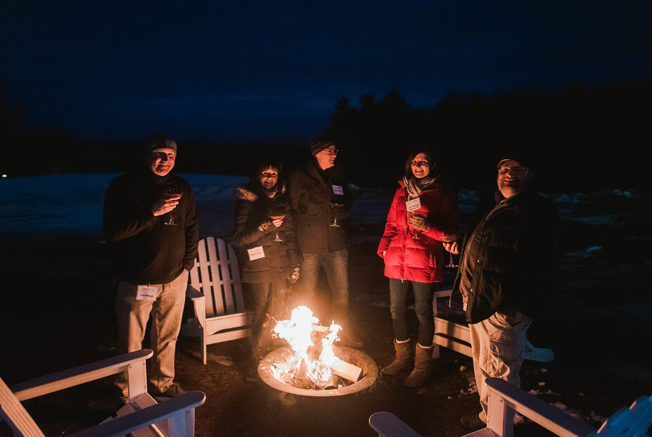 A group of parents stands around an outdoor fire pit at night, holding drinks. They are dressed warmly and surrounded by redesigned Adirondack chairs. Saint Joseph's College of Maine