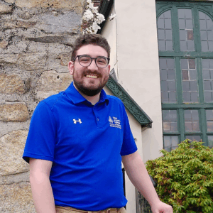 A man wearing glasses and a blue polo shirt stands outdoors near a stone wall and a building with green window frames, possibly during an Admissions On Campus event. Saint Joseph's College of Maine