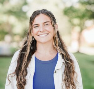 A young woman with long wavy hair, wearing a blue top and light jacket, smiles at the camera outdoors with greenery in the background. Saint Joseph's College of Maine