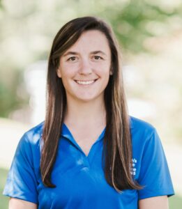A person with long dark hair wearing a blue collared shirt stands outside, smiling at the camera. Saint Joseph's College of Maine