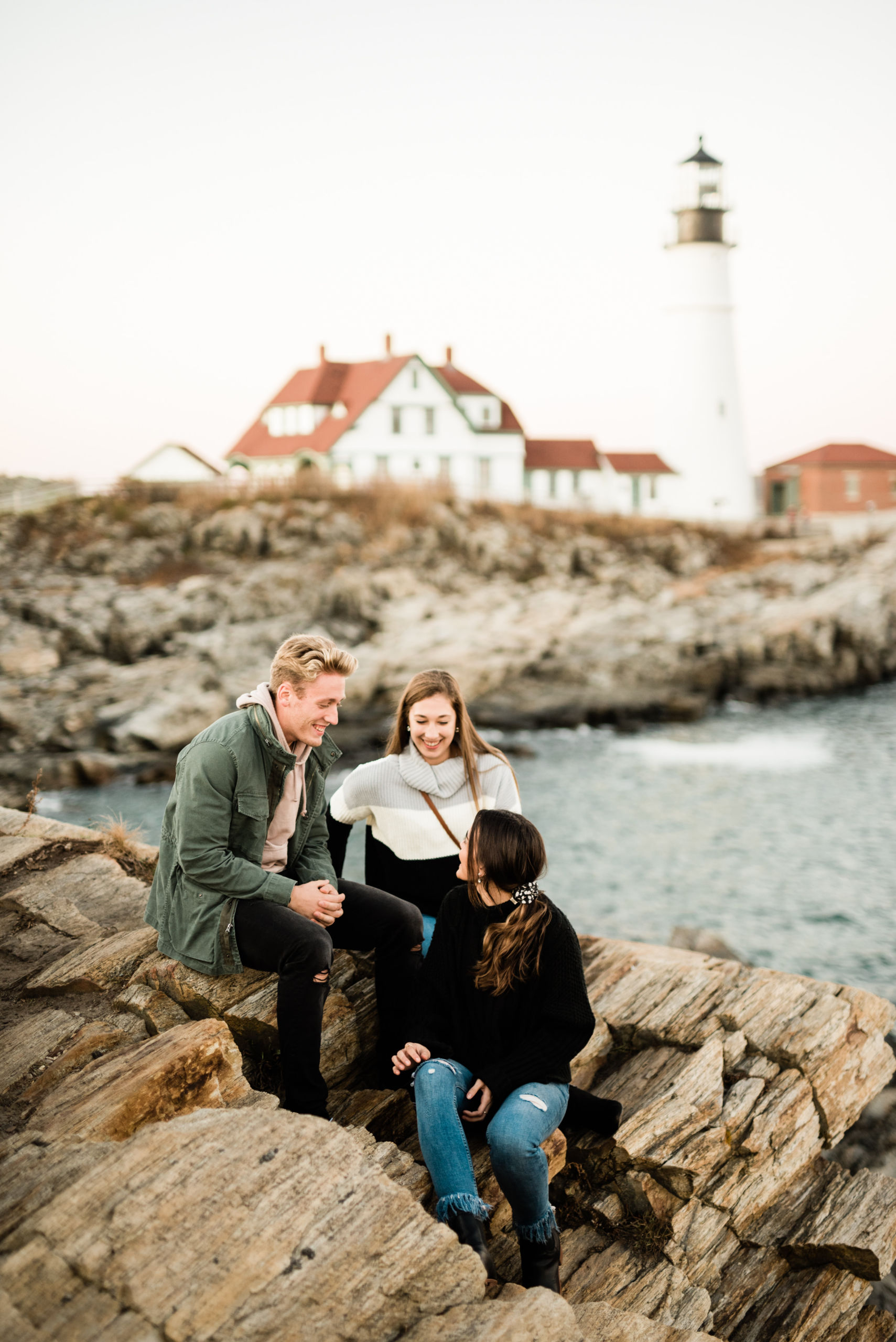 Three people sit on rocky terrain near a shoreline, with a lighthouse and houses visible in the background. They appear to be engaged in conversation. Saint Joseph's College of Maine