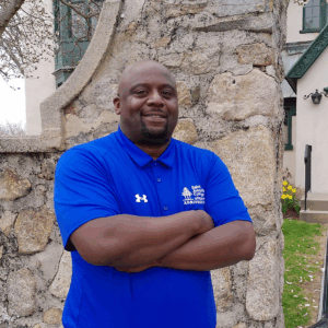 A man in a blue Saint Joseph's College Admissions On Campus polo shirt stands with arms crossed in front of a stone wall and building. Saint Joseph's College of Maine