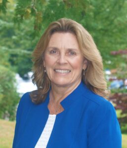 A woman with shoulder-length hair wearing a blue jacket smiles outdoors with greenery in the background. Saint Joseph's College of Maine