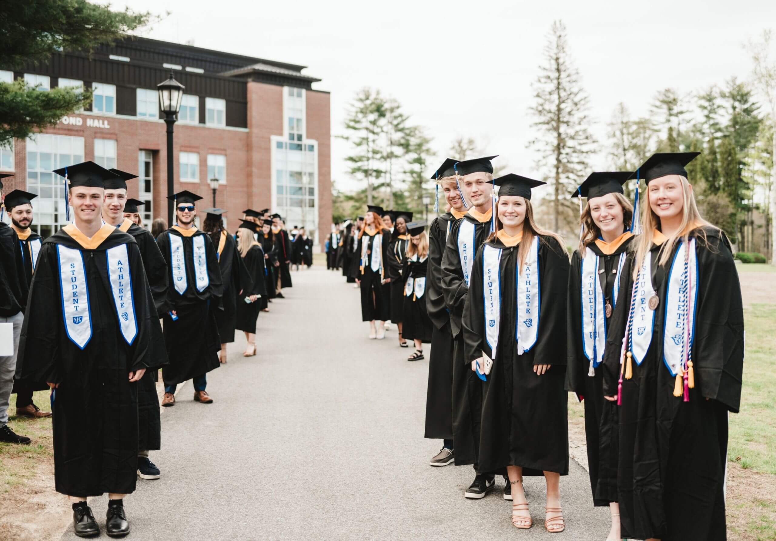 A group of graduating students in caps and gowns stand in two lines on a campus path, celebrating commencement. Some are smiling and holding diplomas, with a building and trees in the background. Saint Joseph's College of Maine