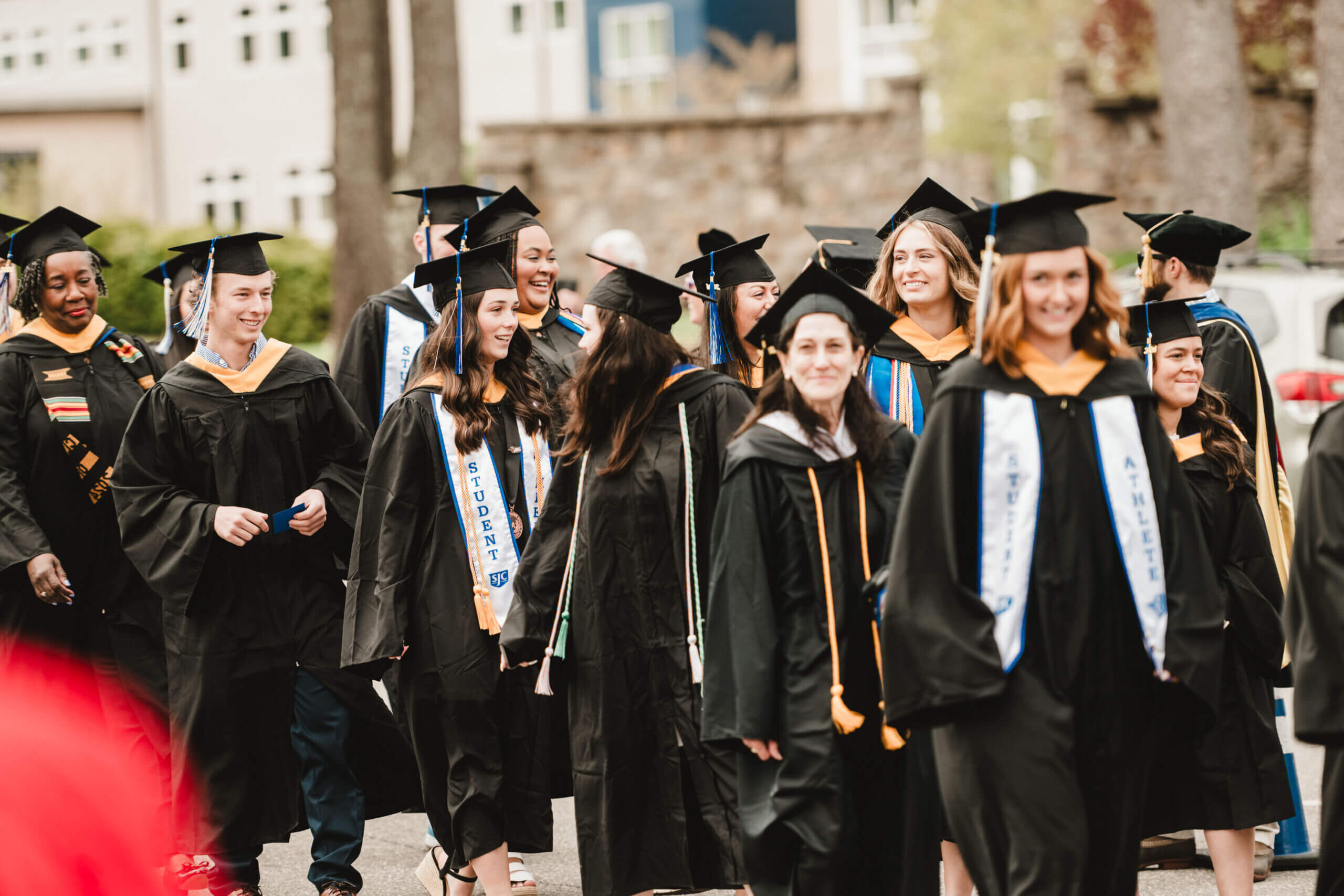 A group of graduates in caps and gowns stroll outdoors, beaming with joy at their commencement. Some proudly wear stoles adorned with text, while the backdrop reveals a blend of architecture and lush greenery. Saint Joseph's College of Maine