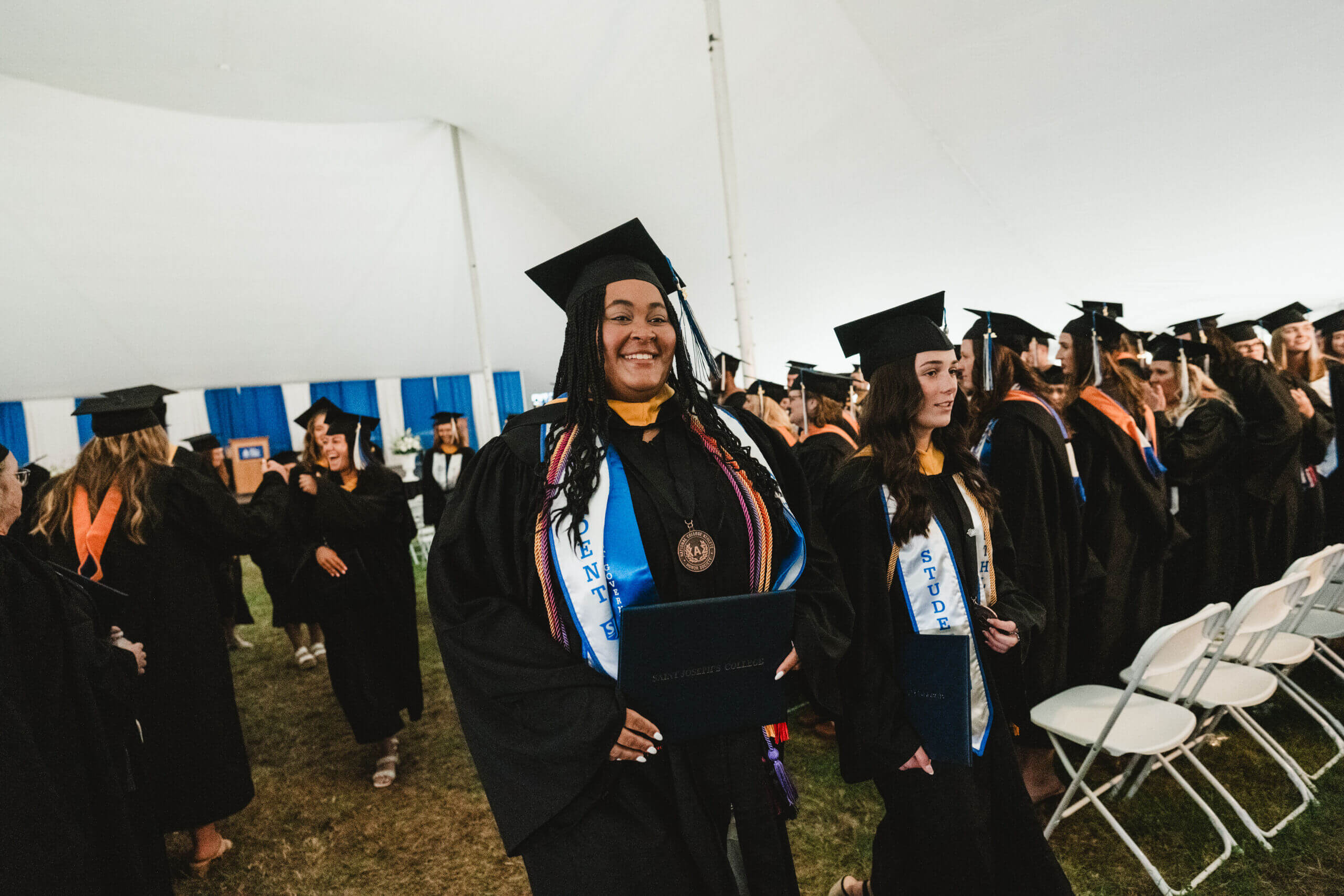 Graduates in caps and gowns beam with pride, holding their diplomas inside the tent during the commencement ceremony. Saint Joseph's College of Maine