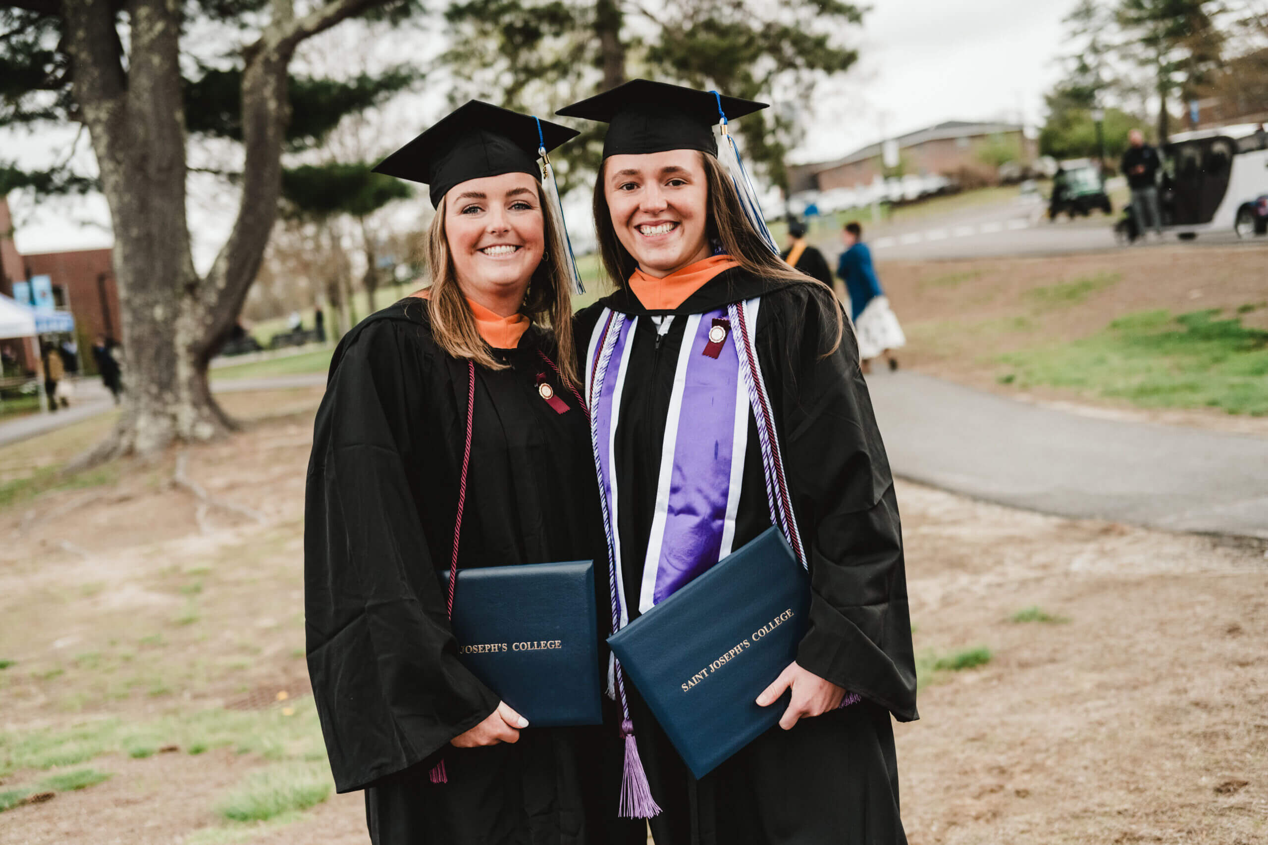 Two graduates in caps and gowns smile brightly during commencement, holding their diplomas while standing outdoors near a path lined with trees. Saint Joseph's College of Maine