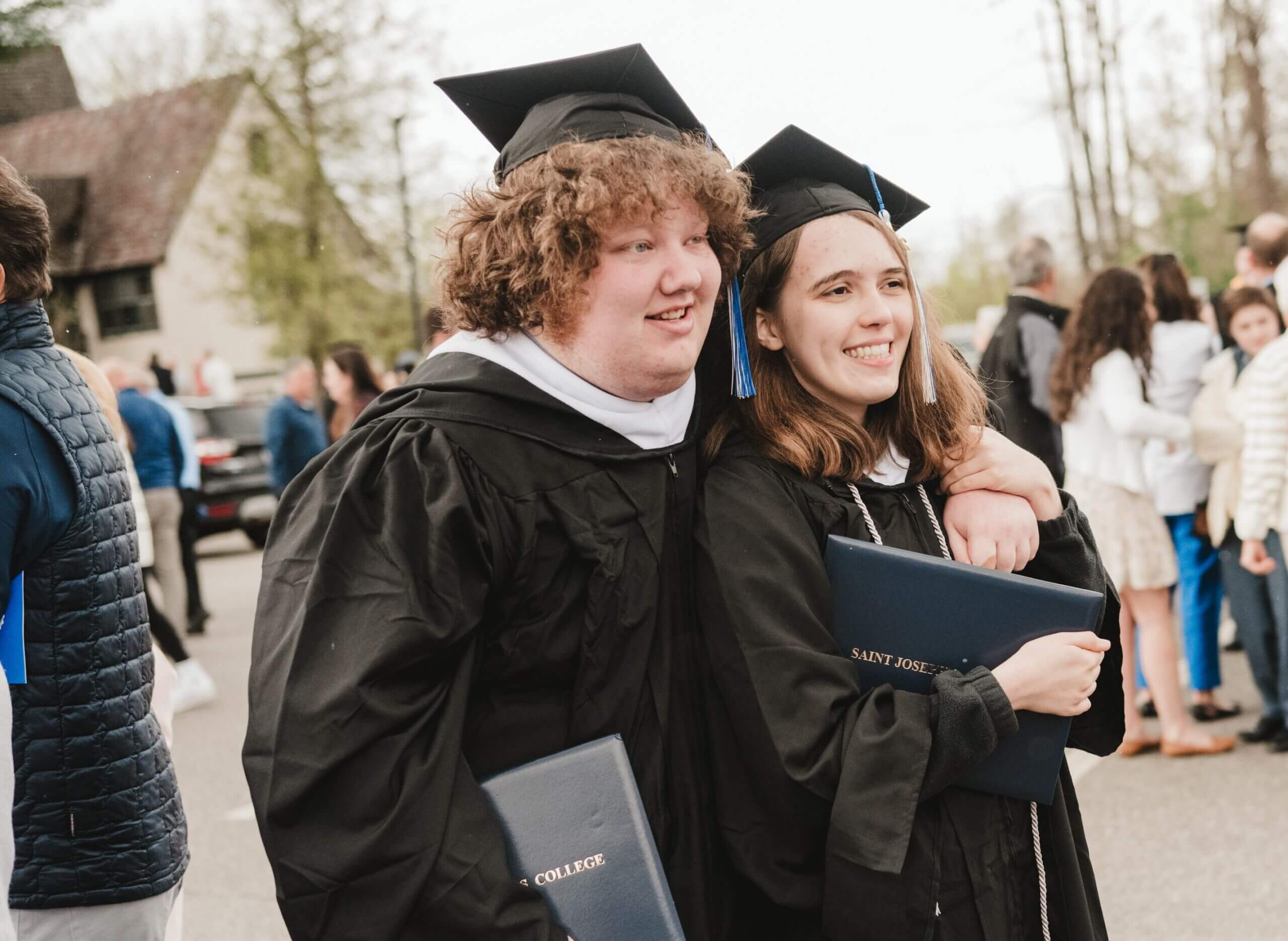 Two people in graduation gowns and caps stand together outdoors, each holding a diploma, basking in the joy of commencement. They appear to be celebrating with others in the background. Saint Joseph's College of Maine