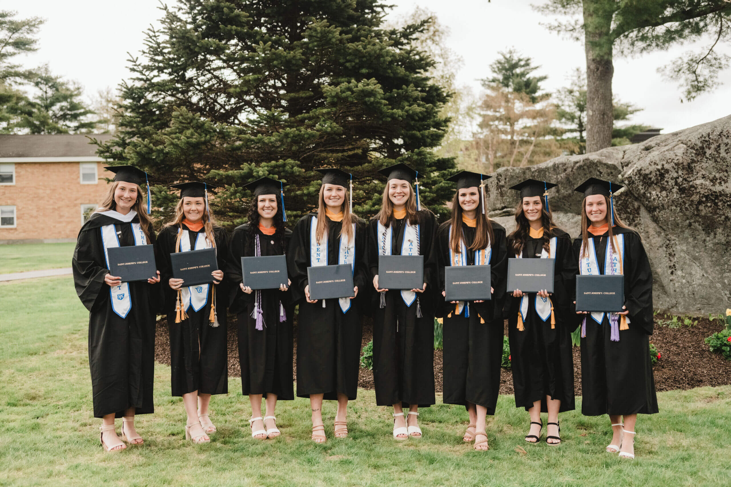 Eight graduates in black gowns and caps celebrate commencement by holding diplomas as they stand on a lush lawn, framed by trees and rocks. Saint Joseph's College of Maine