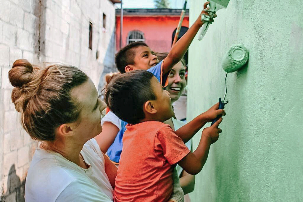Three adults and two children are painting a wall green together. One child uses a roller while another holds a paintbrush. They appear to be in an outdoor setting. Saint Joseph's College of Maine